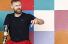 A white man with brown hair, a brown beard and a prosthetic arm is sat down. 